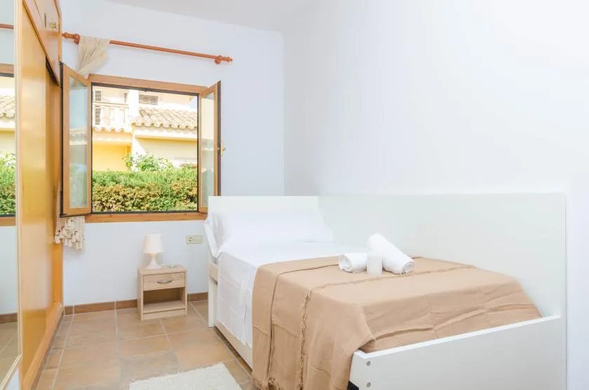 A bright bedroom with white bedding and an open window overlooking greenery.