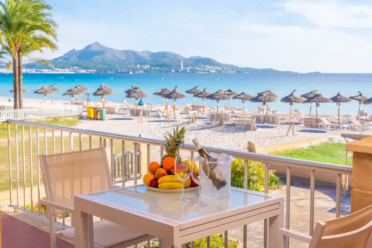 Una terraza junto al mar da a una playa de arena, aguas turquesas y montañas lejanas.