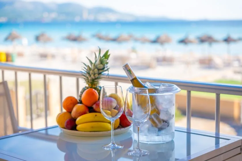 Una mesa de balcón junto al mar tiene fruta, copas de vino y una botella fría.