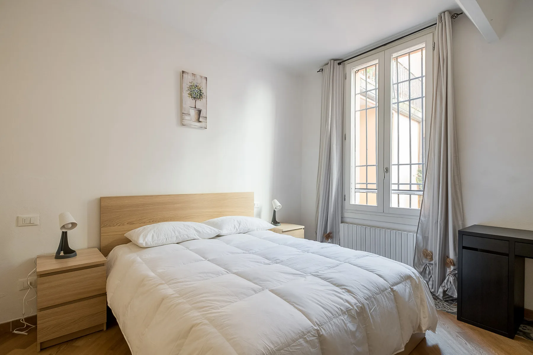 A bright bedroom with a neatly made bed beside tall windows.
