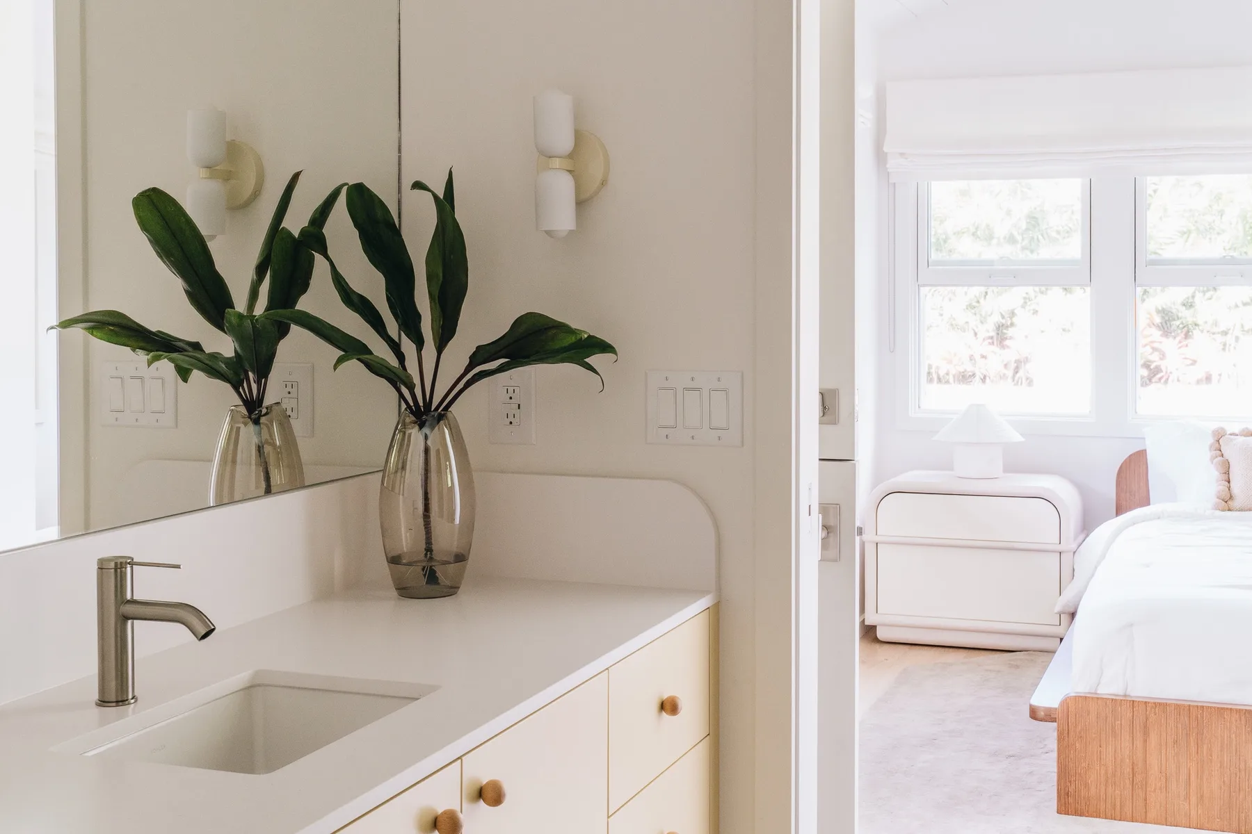 A bright bedroom with a bed beside a vanity and leafy plants.