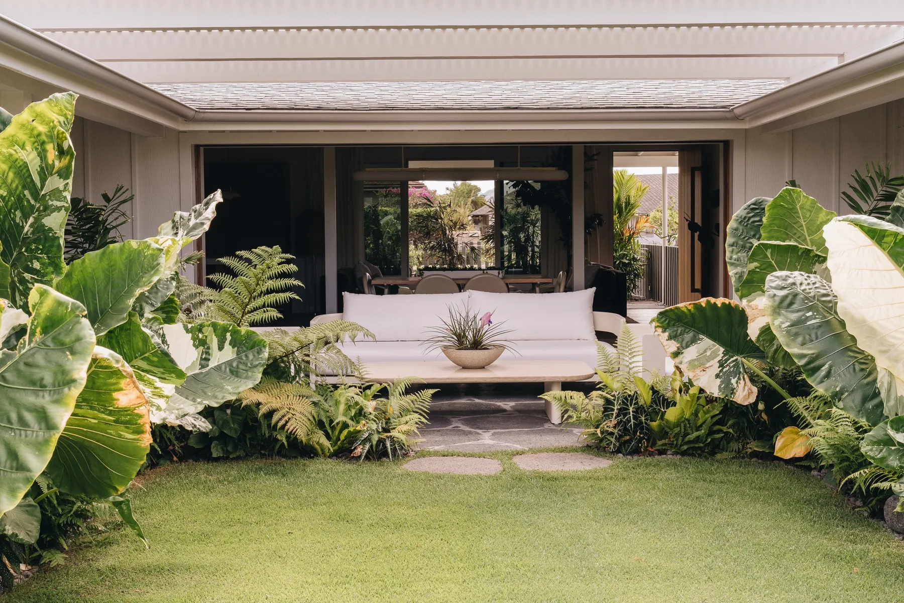 A covered garden seating area opens onto a lawn, with plants surrounding the sofa.