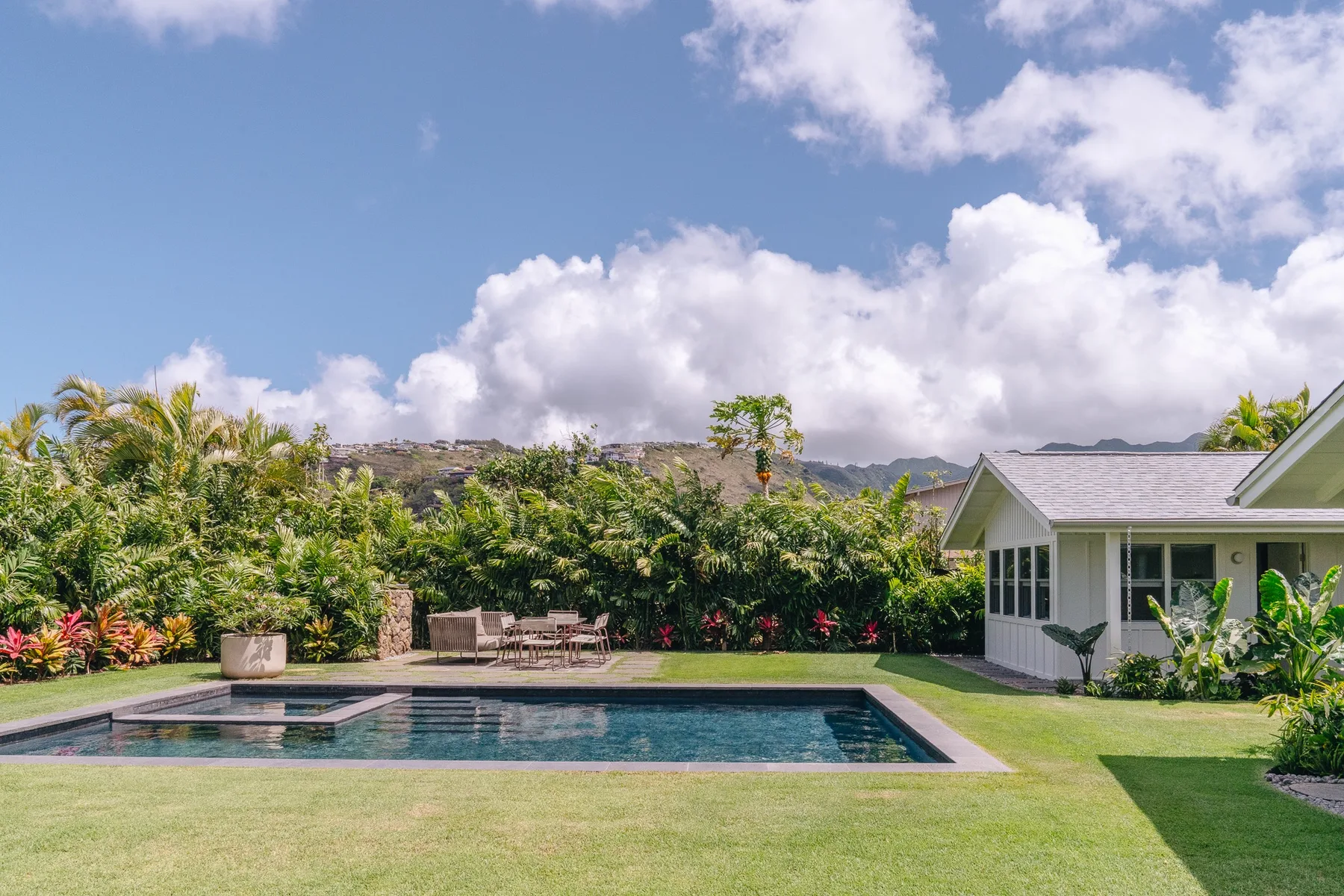 An outdoor swimming pool sits beside a small light-colored building and landscaped greenery.