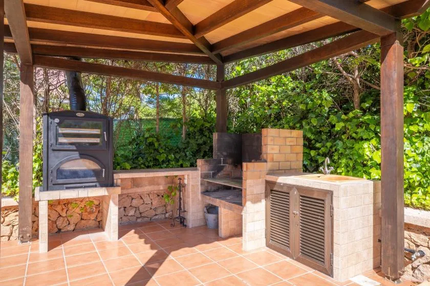 Una zona de cocina al aire libre bajo una pérgola de madera, con horno de ladrillo y encimera de azulejos.