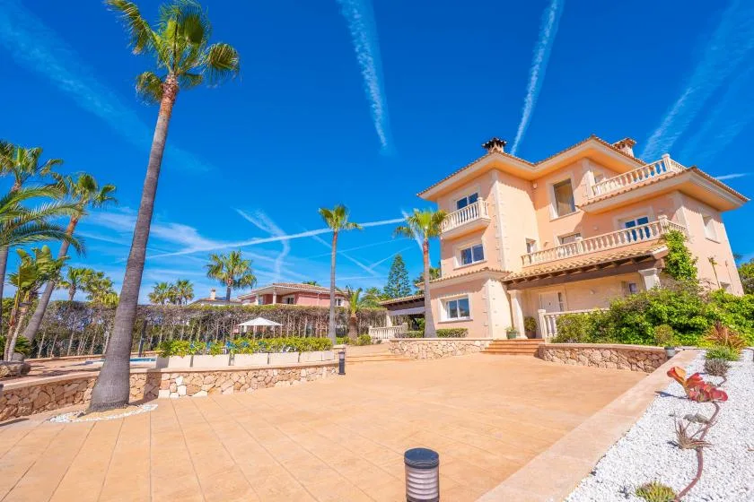 Una villa color melocotón se alza junto a un amplio patio pavimentado con palmeras.