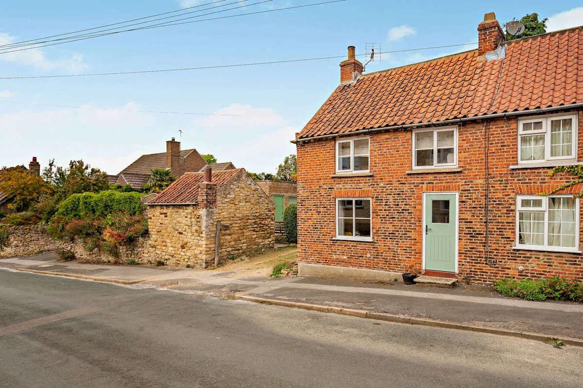 A brick cottage with a tiled roof stands beside a stone garden wall along the street.