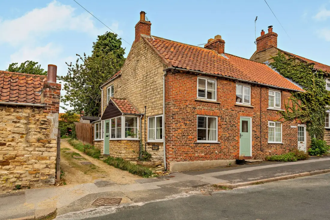 A brick house stands beside a narrow lane, with a tiled roof and small garden.