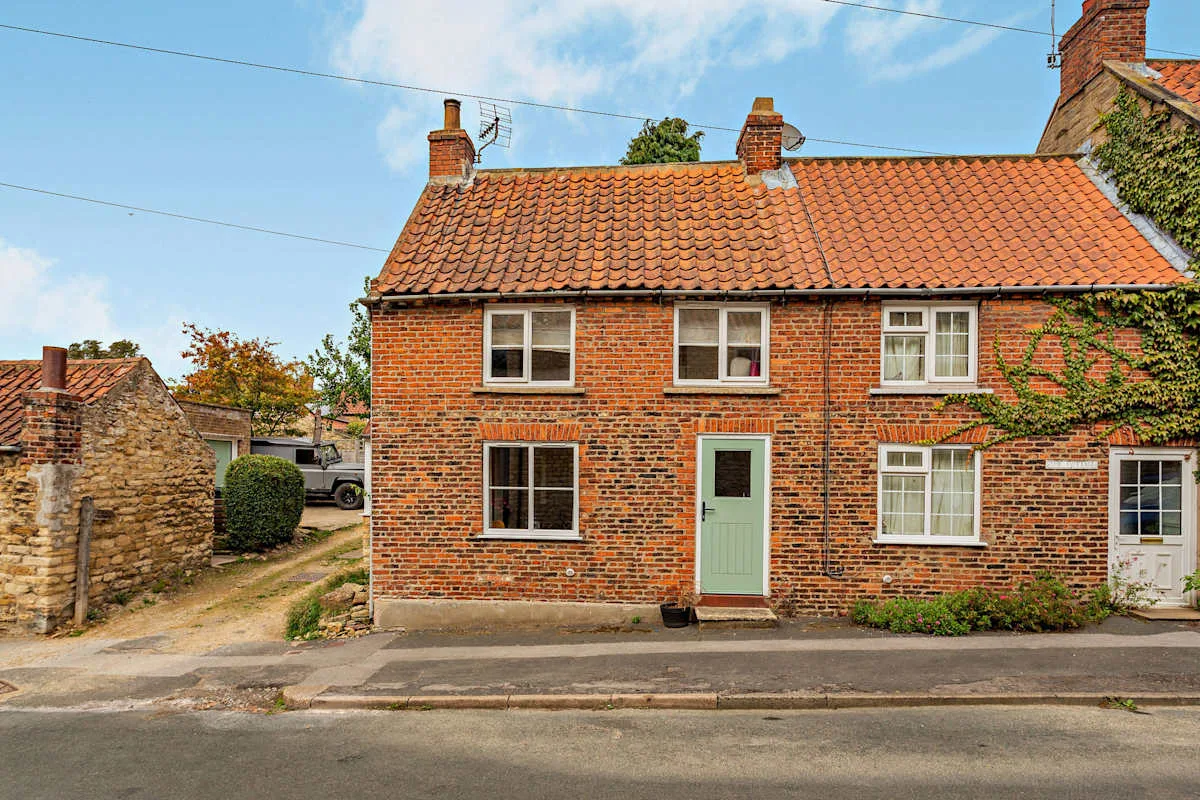 A red-brick cottage stands beside a road, with a tiled roof and pale green door.
