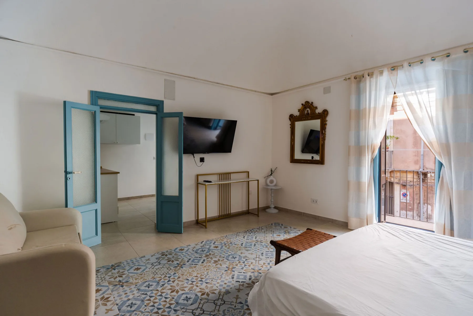 Un dormitorio con cama, asientos y una alfombra estampada junto a una puerta abierta al balcón.