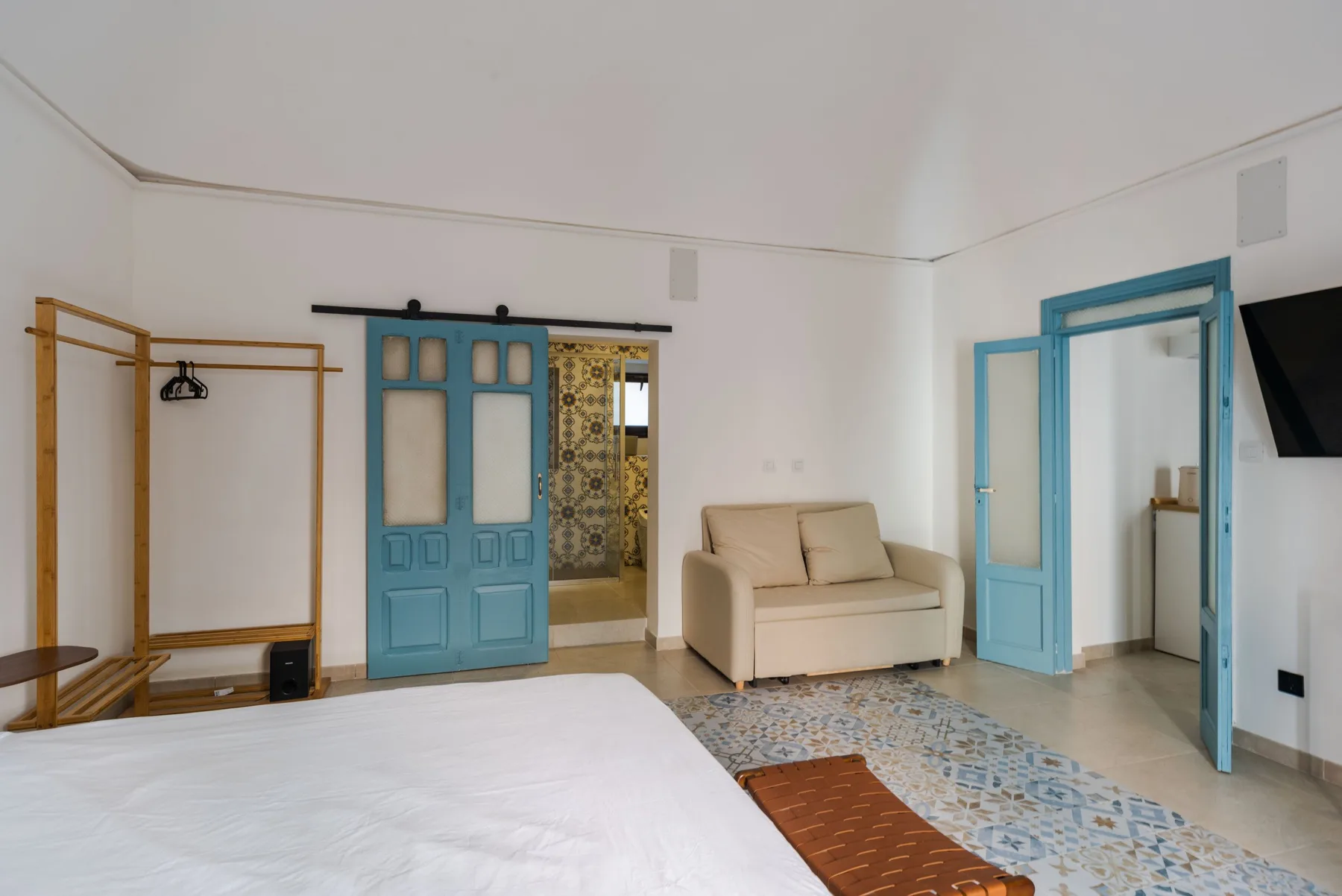 Una habitación luminosa con cama, asientos beige y puertas azules que dan a una zona alicatada.