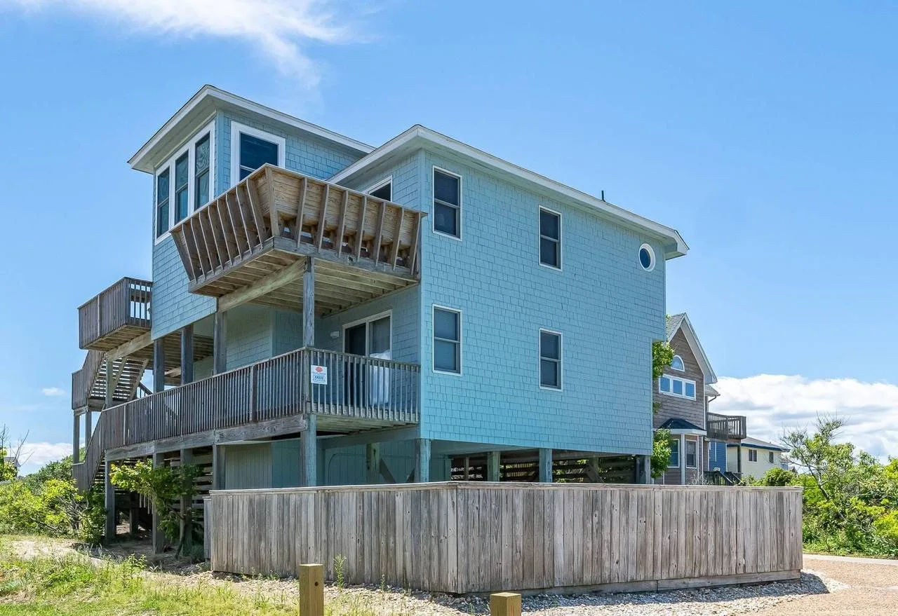 Una casa azul elevada con balcones de madera está junto a un patio cercado.