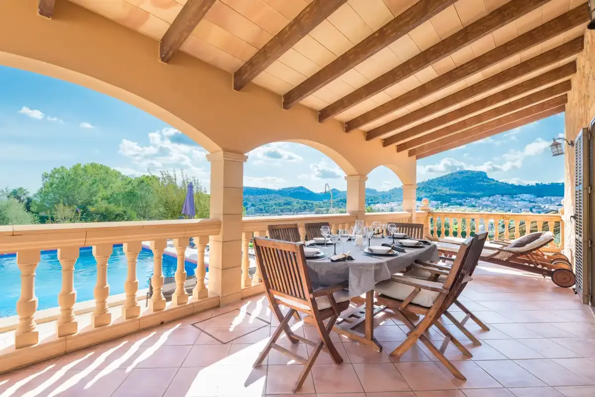A covered terrace with a dining table and seating overlooks a pool and distant hills.