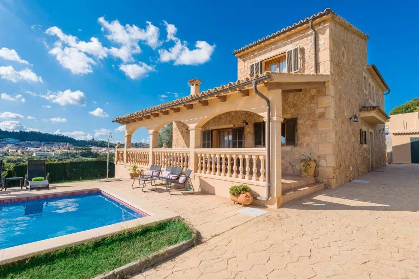 A stone house with a covered patio and swimming pool under a bright blue sky.