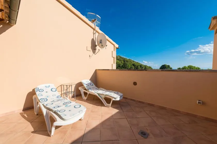A sunlit terrace with reclining seating and a low wall beneath a blue sky.