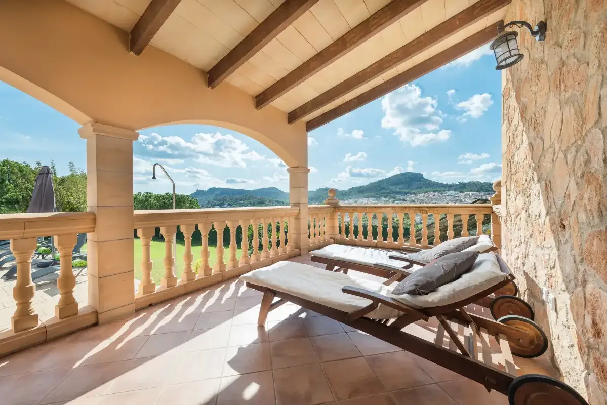 A covered terrace has cushioned loungers overlooking a green landscape and hills.