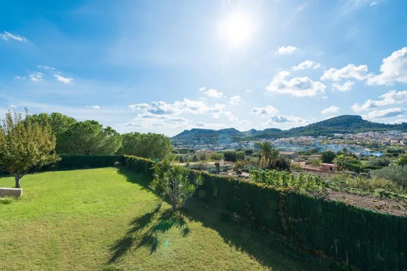 A sunny garden lawn bordered by hedges overlooks a distant town and hills.