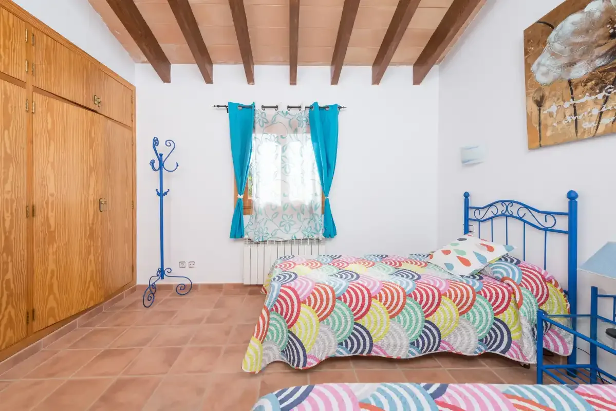A bedroom with a blue metal bed, colorful patterned bedding, and a curtained window.