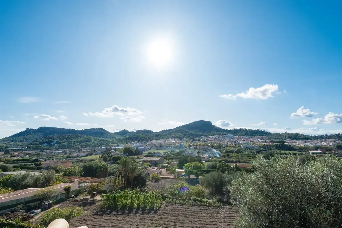 A sunny rural landscape with fields, scattered homes, and hills in the distance.