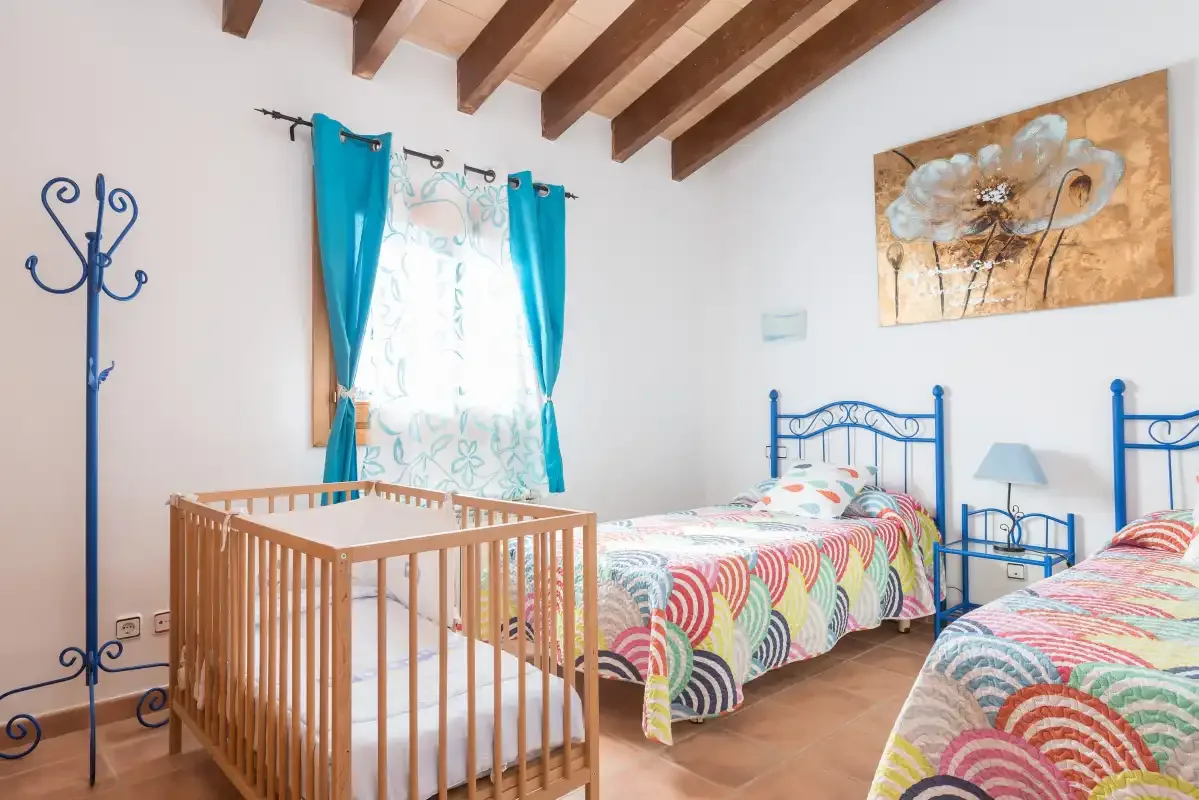 A bright bedroom with colorful beds and a wooden crib beside the window.