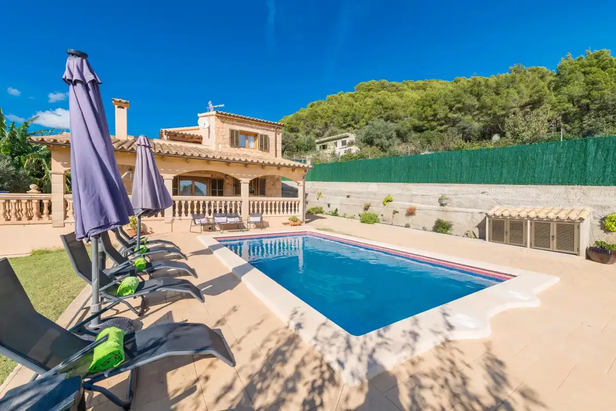 A swimming pool beside a house, with loungers and umbrellas on the patio.