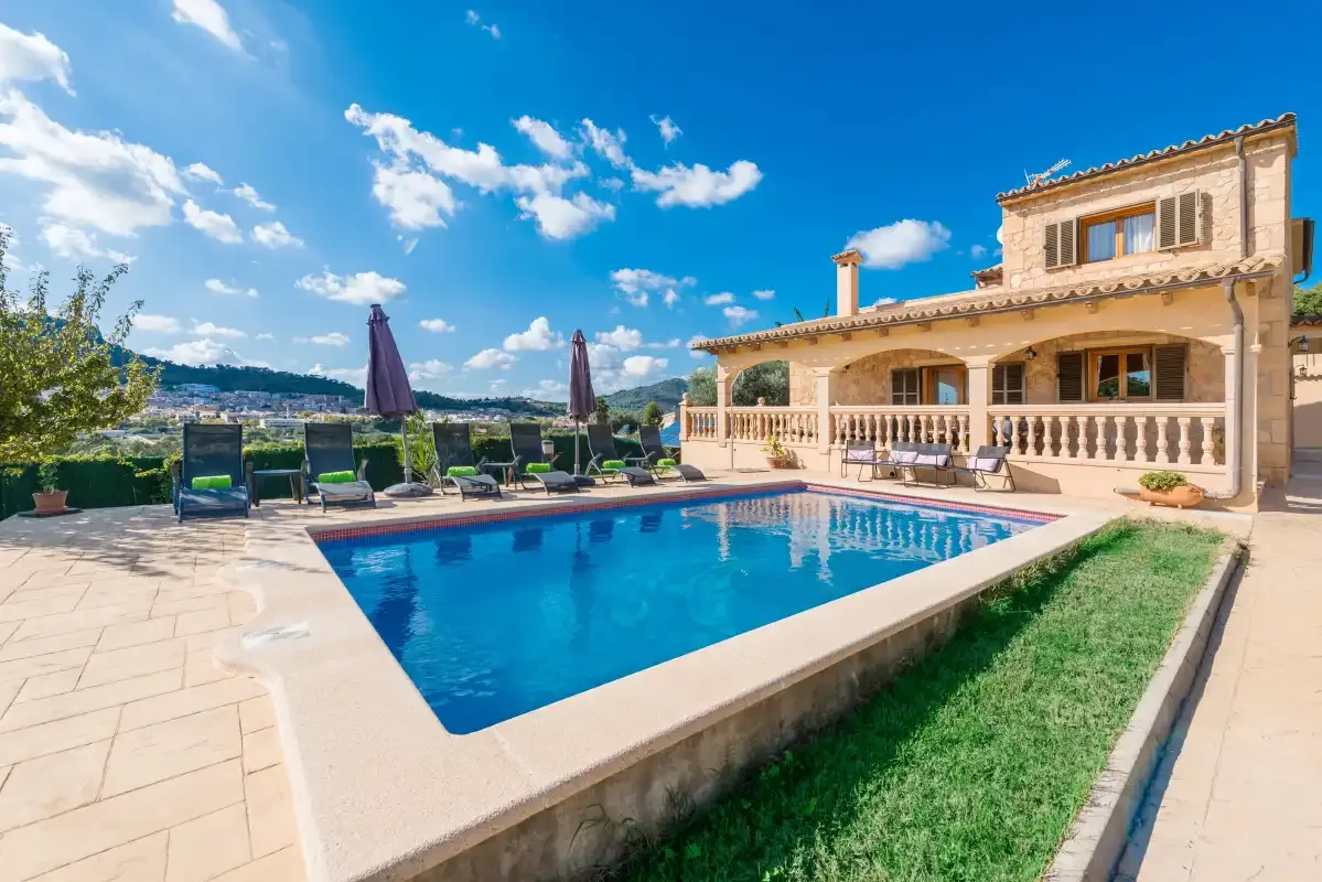 A swimming pool sits beside a stone house and a paved patio.
