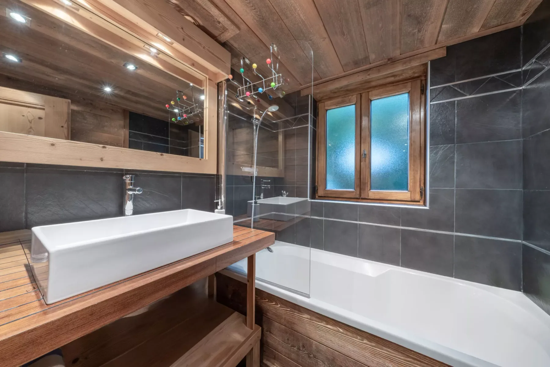 Un baño con mueble de madera, lavabo sobrepuesto y bañera bajo una ventana.