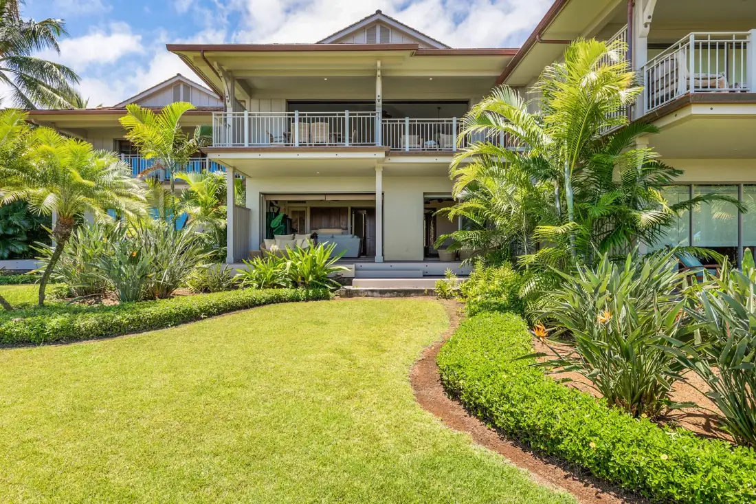 A two-story house faces a green lawn, with tropical plants along the garden edges.