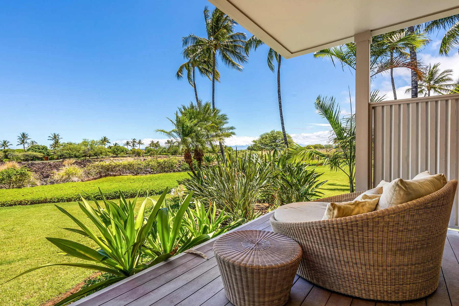 A covered patio with woven seating overlooks a green lawn and tropical plants.