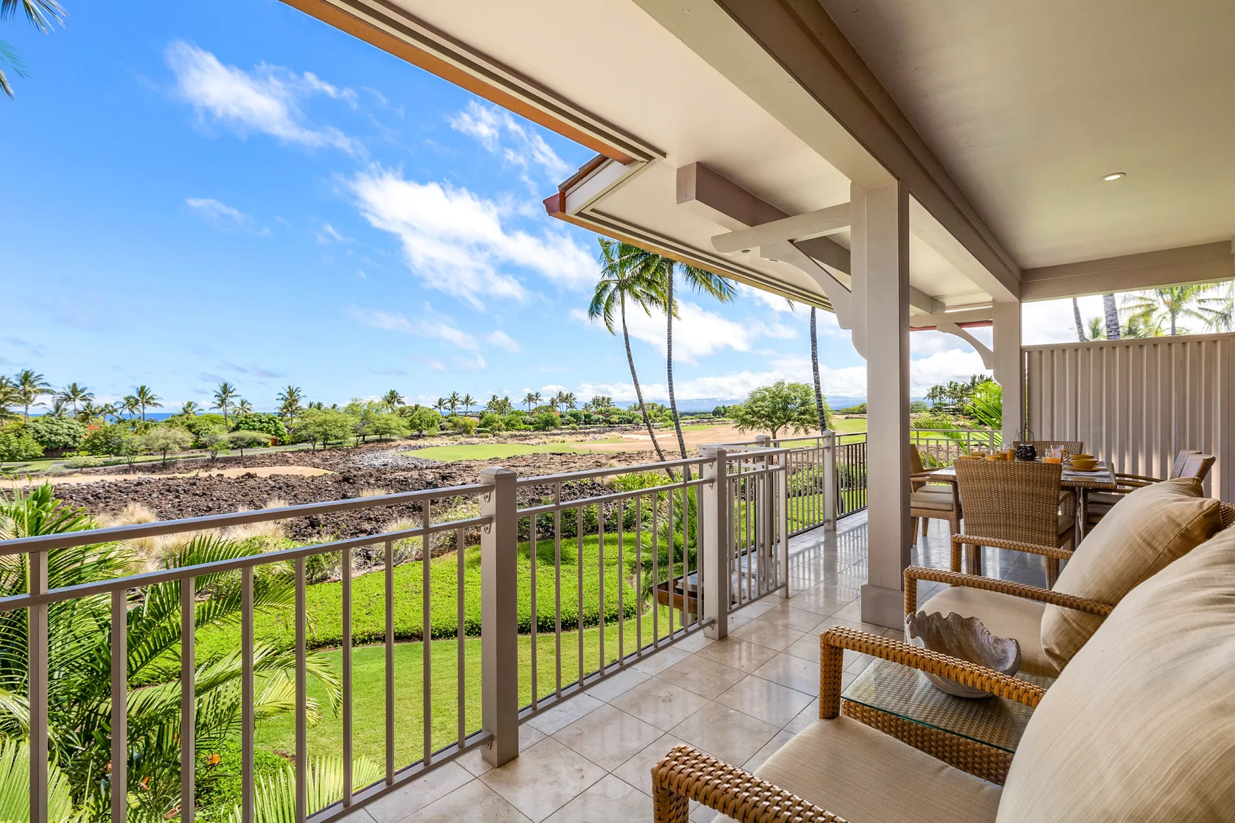 A covered patio with wicker seating overlooks a green landscaped area and distant ocean.