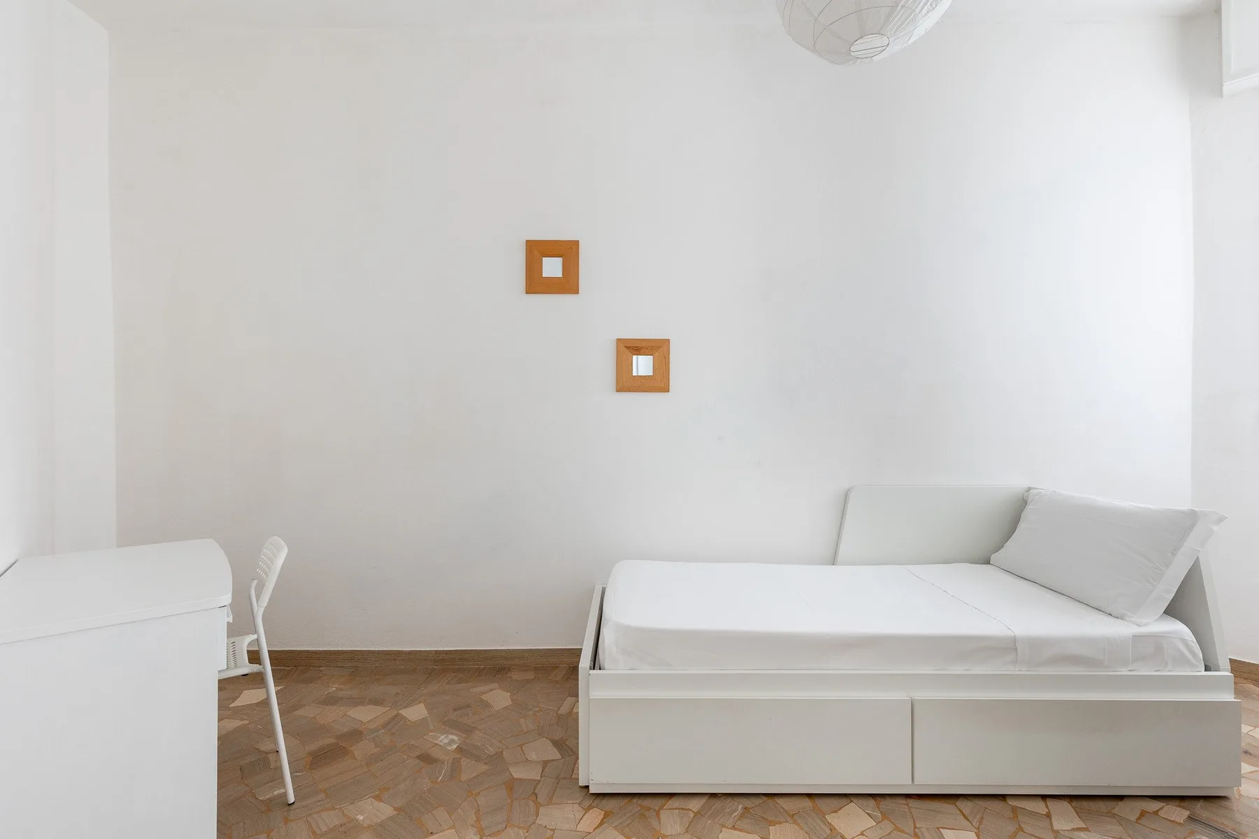 Una habitación luminosa y blanca con una cama de día, un escritorio y dos cuadros pequeños.