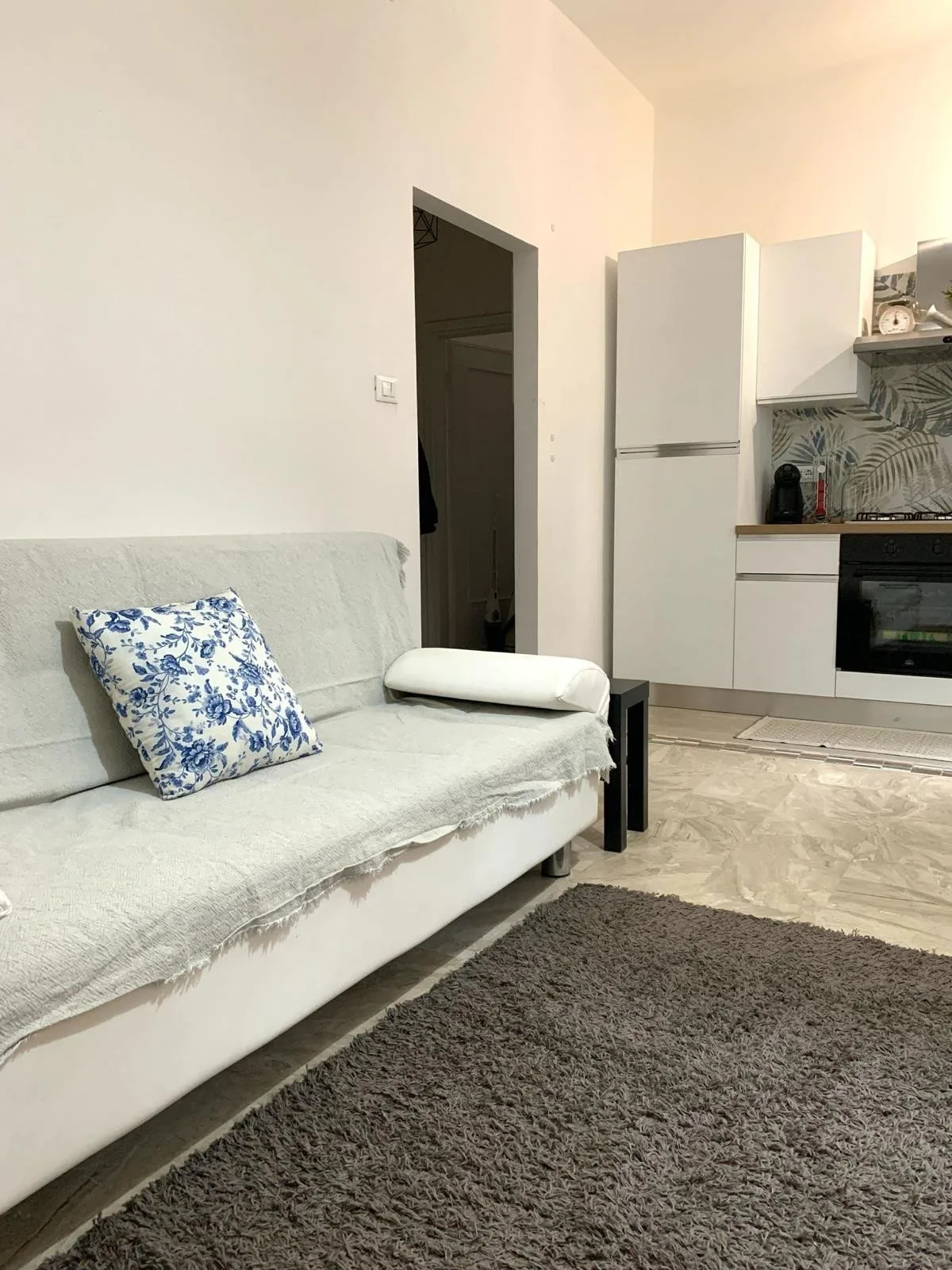 Una sala de estar con asientos claros, un cojín estampado y una pequeña zona de cocina.