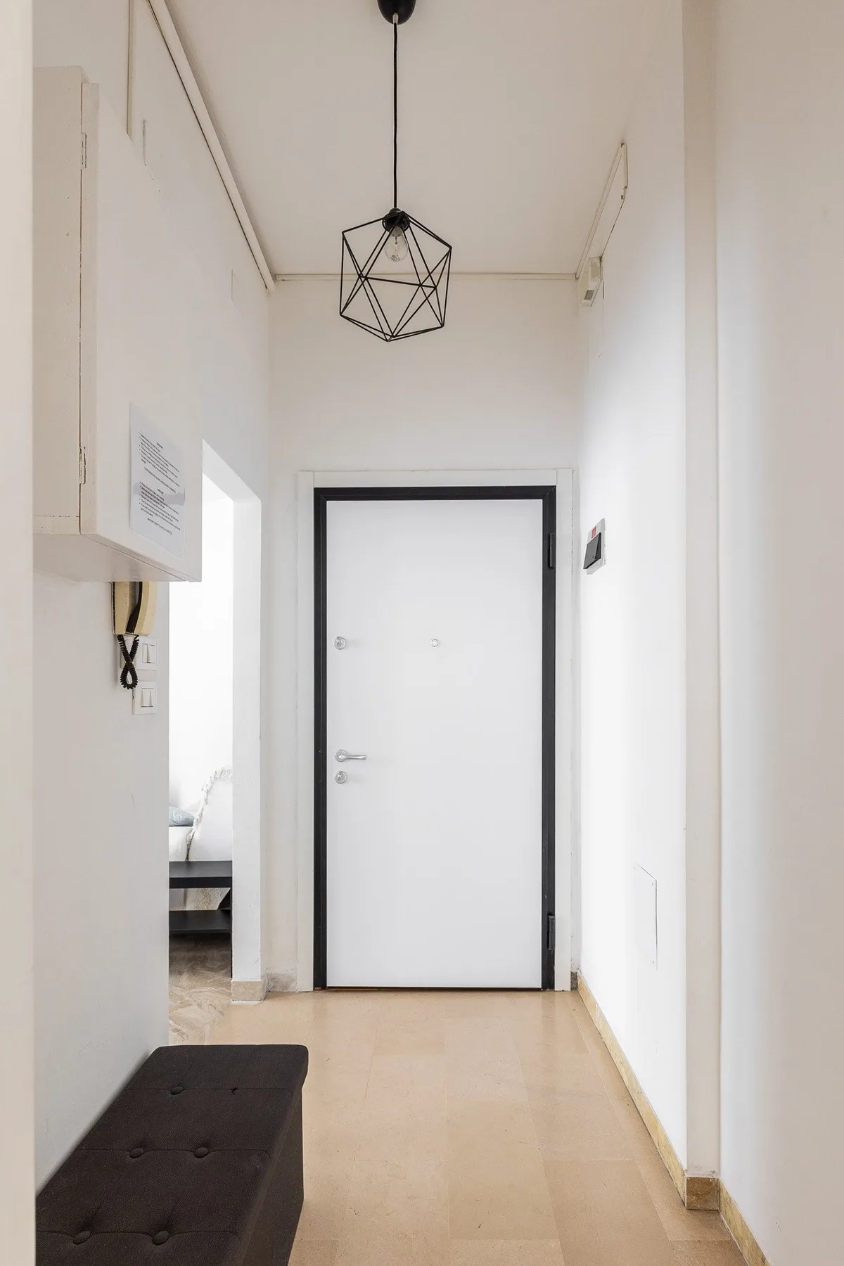 Un pasillo estrecho y blanco con una puerta cerrada y un banco oscuro.