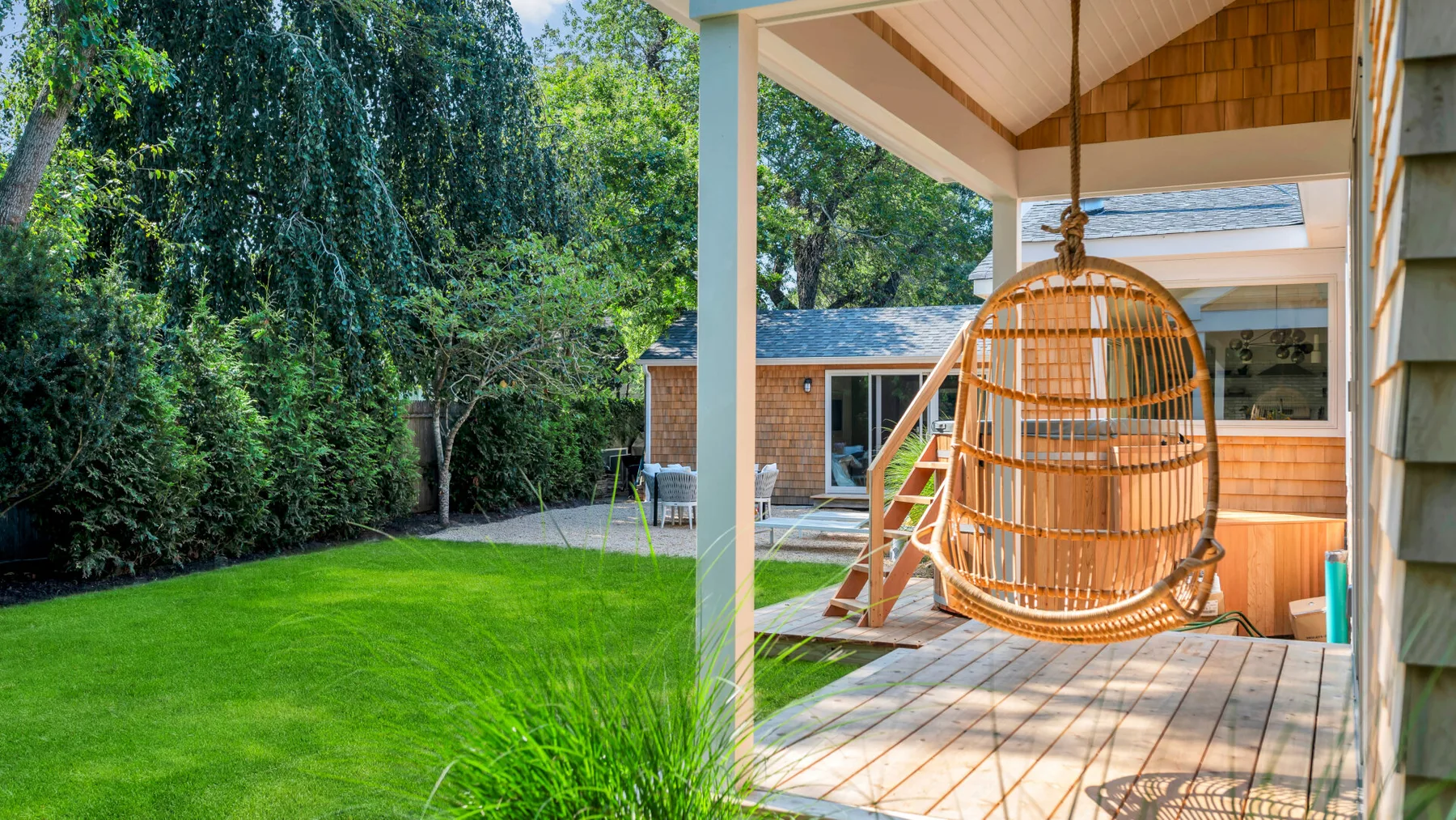 Una terraza de madera cubierta con una silla colgante da a un césped verde y un jardín.