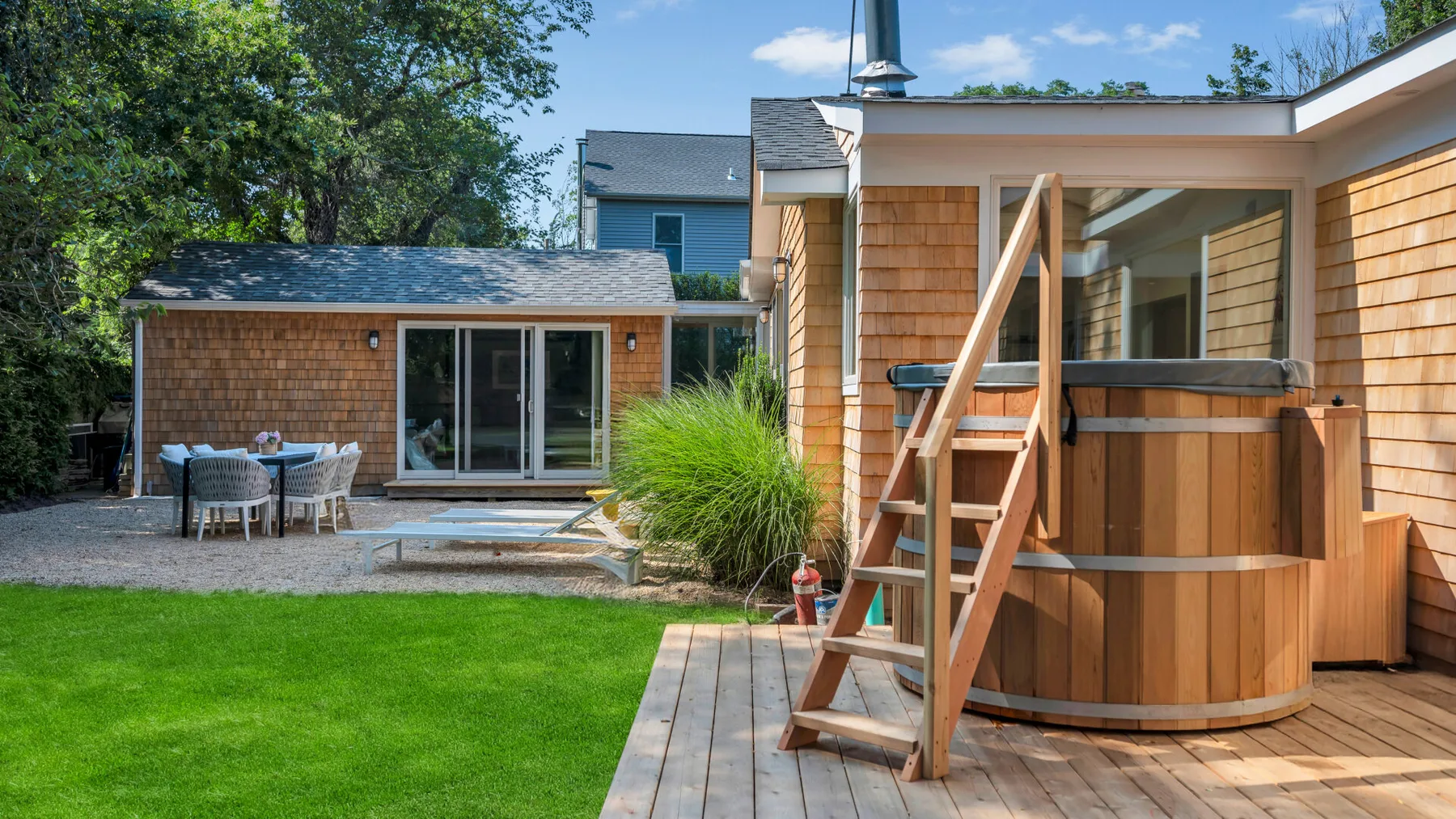 A backyard patio features a wooden hot tub beside a small shingled building.