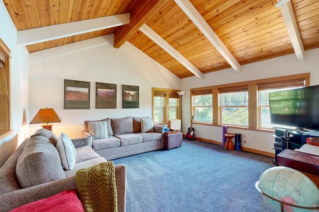 A living room with seating, a television, and large windows beneath a wood-beamed ceiling.