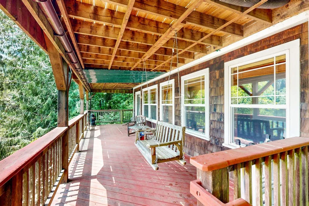 Una terraza de madera cubierta, con barandilla y ventanas a un lado.