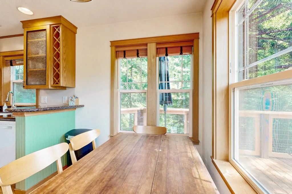 Un comedor con una mesa de madera junto a grandes ventanas que dan a la vegetación.