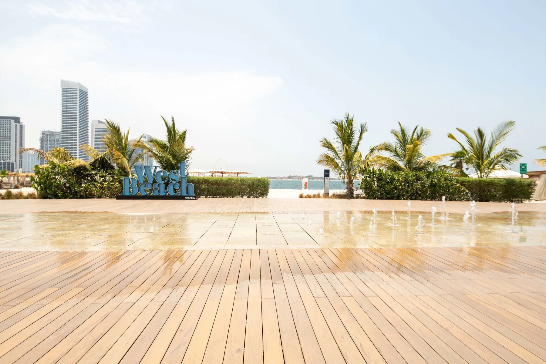 Un paseo junto al agua con tarima de madera, palmeras y edificios al otro lado.