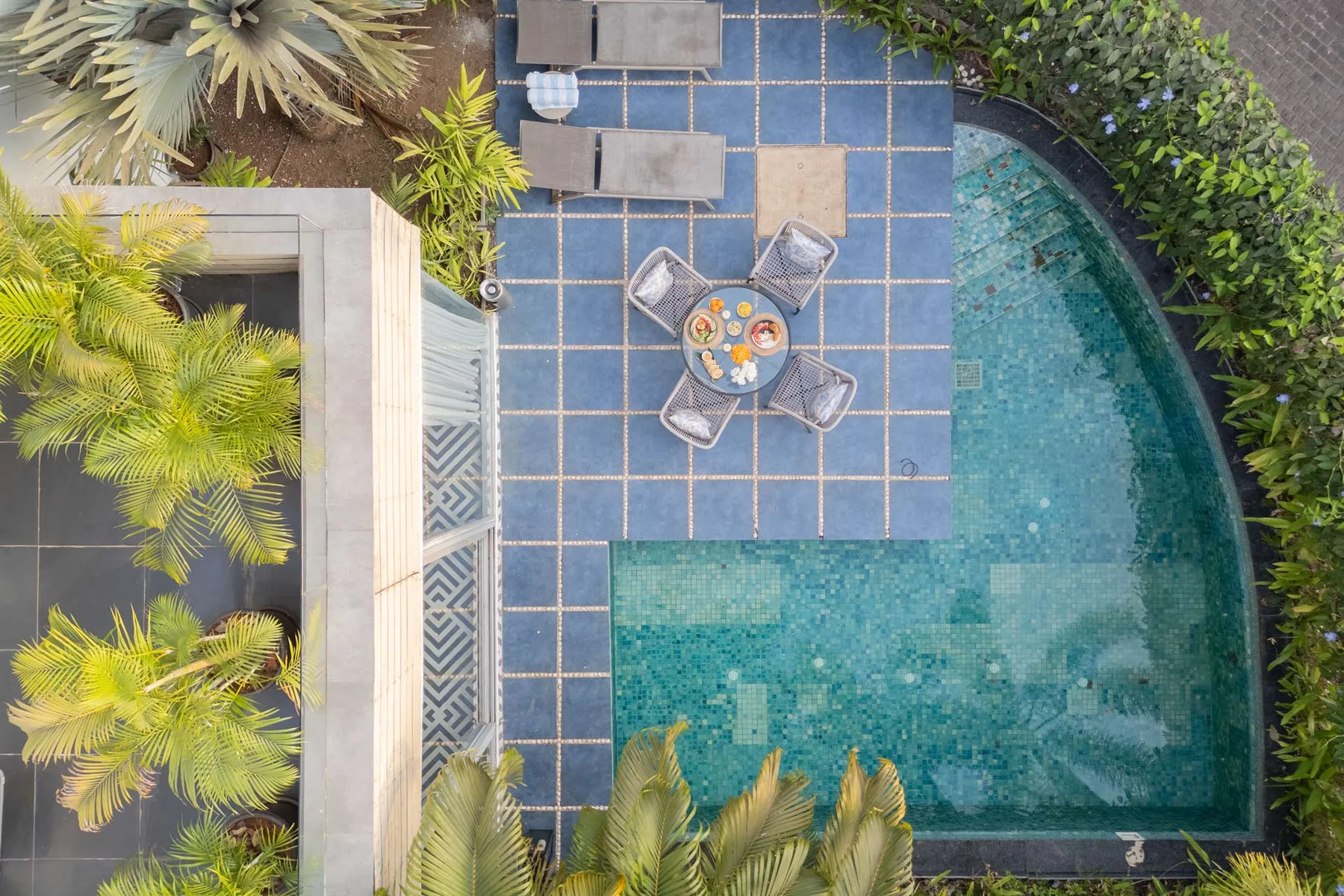 Una piscina al aire libre junto a un patio embaldosado con asientos y plantas alrededor.