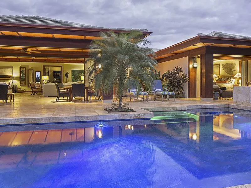 A lit patio with seating and palm trees overlooks a swimming pool at dusk.