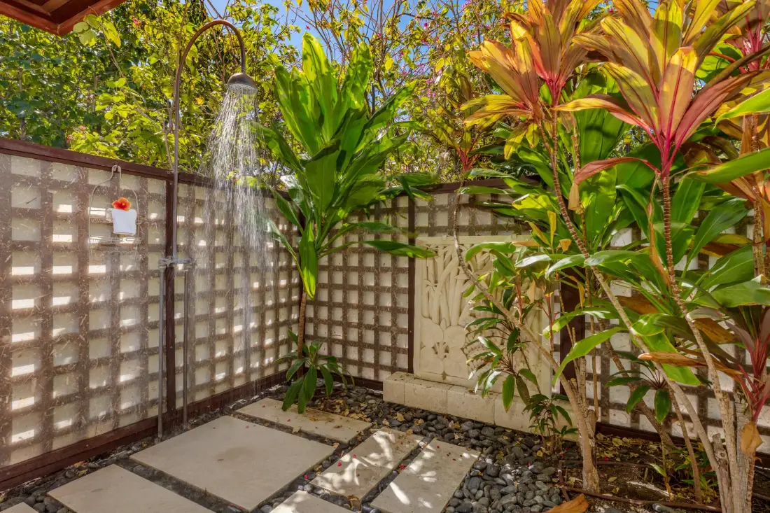 An outdoor shower stands beside a paved garden path and leafy plants.