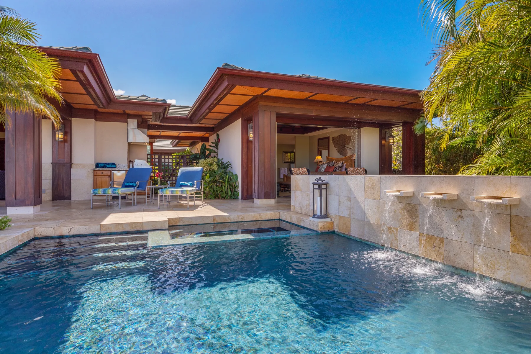 An outdoor pool beside a covered patio with seating and tropical plants.