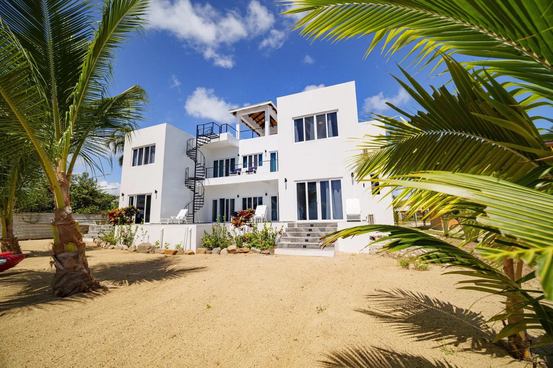 A white modern building stands among palm trees in a sandy outdoor area.