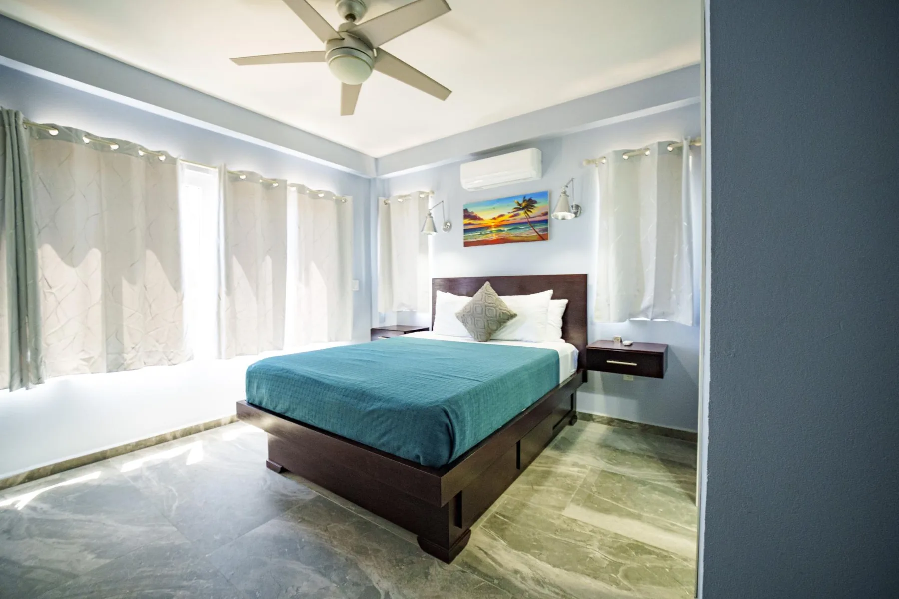 A bedroom with a bed covered in teal bedding and windows with light curtains.