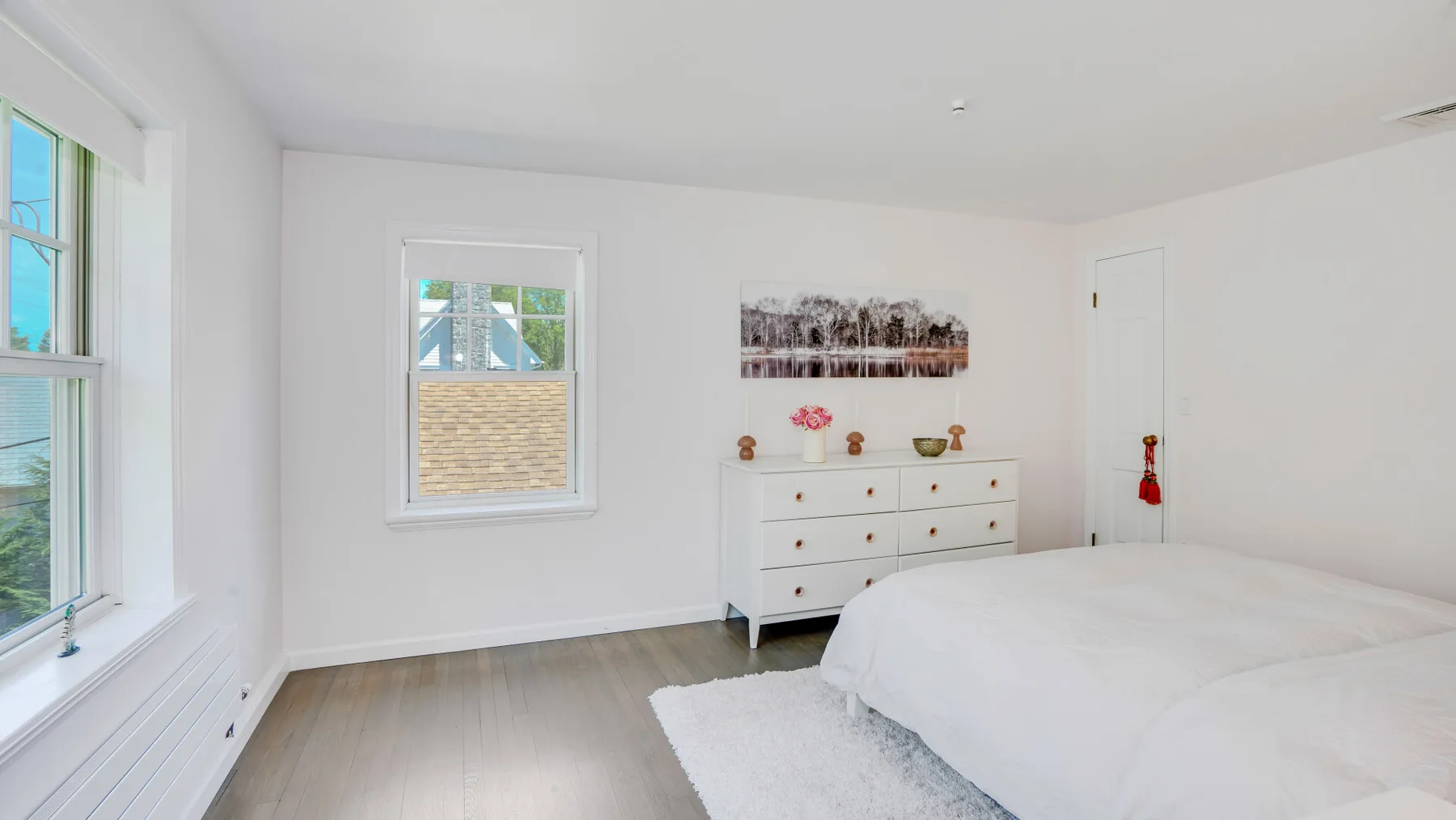 Un dormitorio luminoso con una cama blanca, una cómoda y ventanas grandes.