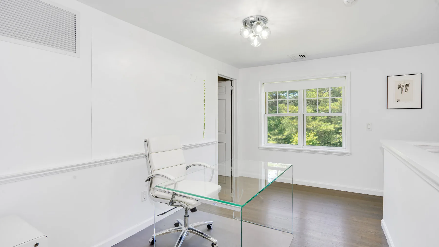 Una habitación luminosa y blanca con un escritorio de vidrio y una ventana amplia con vista a la vegetación.
