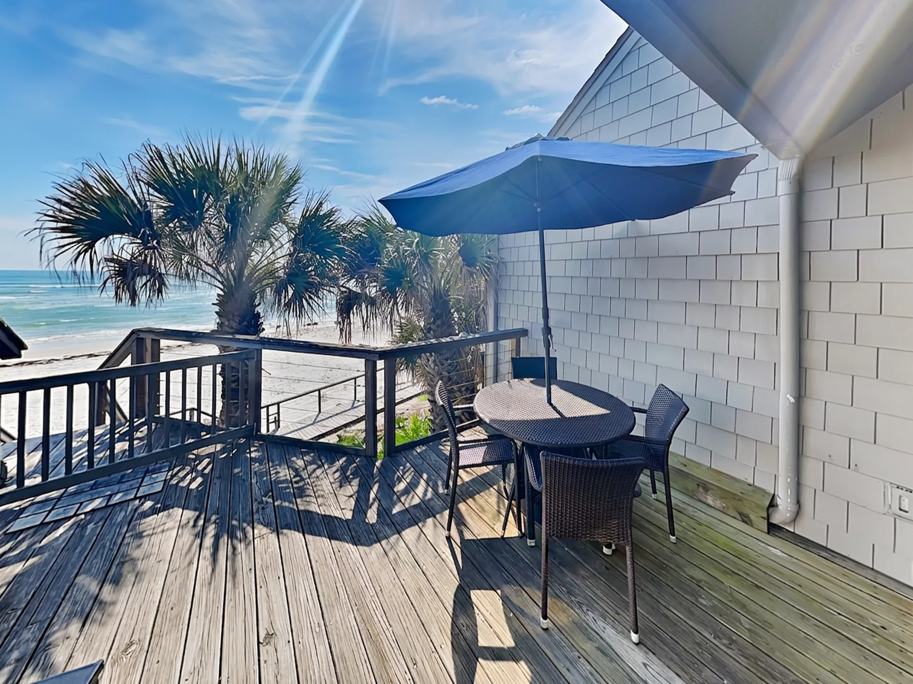 A seaside deck with outdoor seating beneath a blue umbrella, overlooking the ocean.
