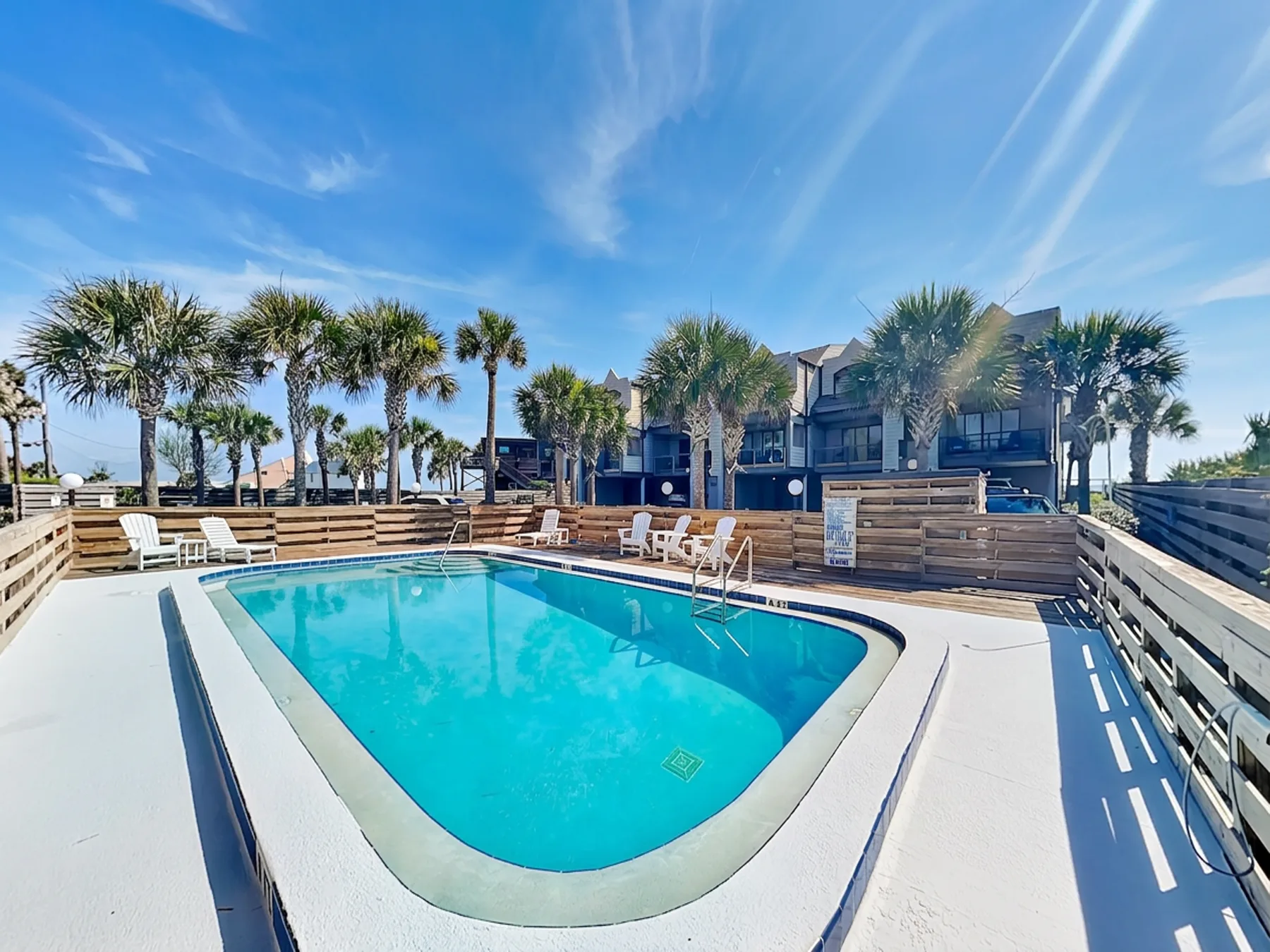 An outdoor swimming pool is surrounded by a deck, railings, and palm trees.