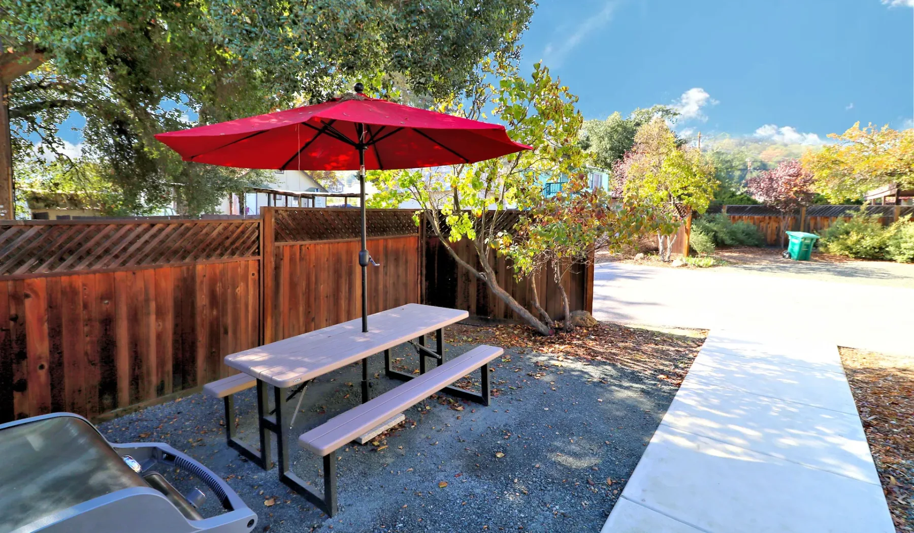 Un patio al aire libre con una mesa de picnic bajo una sombrilla roja junto a una cerca de madera.