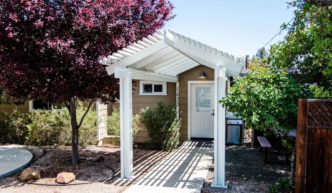 Una pequeña construcción de jardín con puerta blanca está bajo una pérgola.
